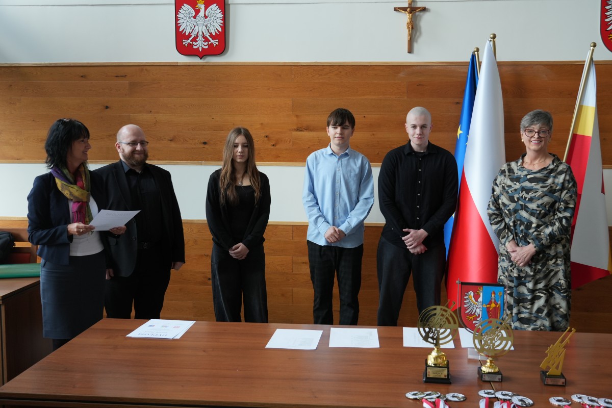 Grupa kilku uczniów pozuje do zdjęcia z dwiema paniami. Uczniowe trzymają dyplomy oraz statuetki.