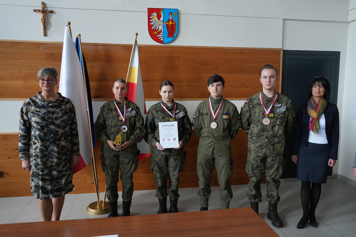 Grupa kilku uczniów pozuje do zdjęcia z dwiema paniami. Uczniowe trzymają dyplomy oraz statuetki.