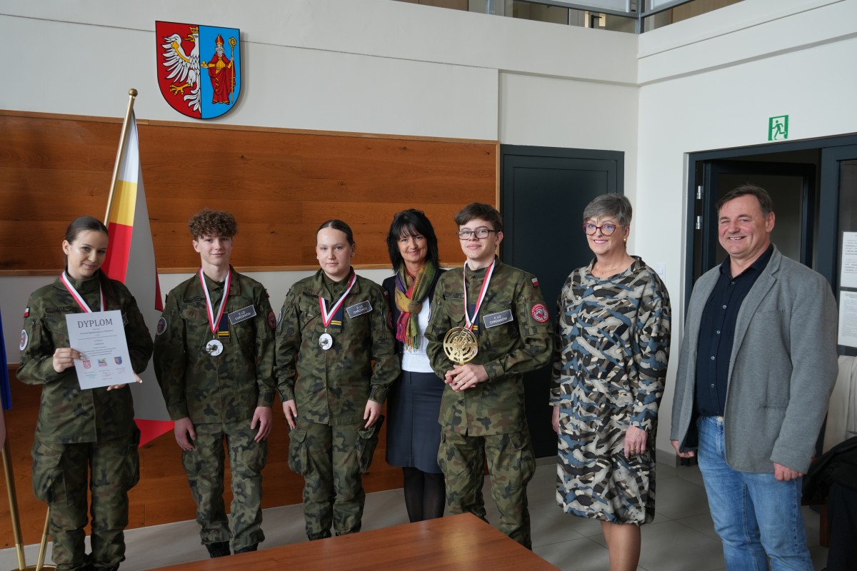 Grupa kilku uczniów pozuje do zdjęcia z dwiema paniami. Uczniowe trzymają dyplomy oraz statuetki.