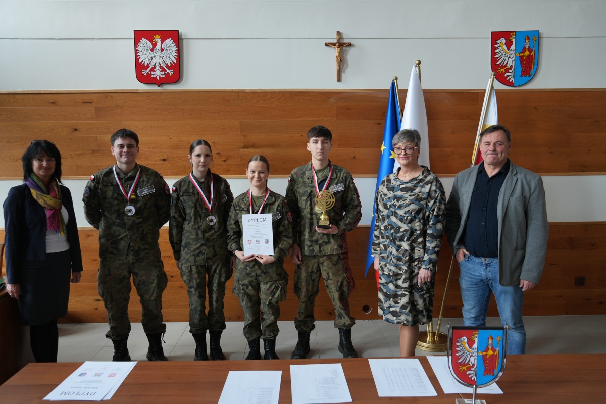 Grupa kilku uczniów pozuje do zdjęcia z dwiema paniami. Uczniowe trzymają dyplomy oraz statuetki.