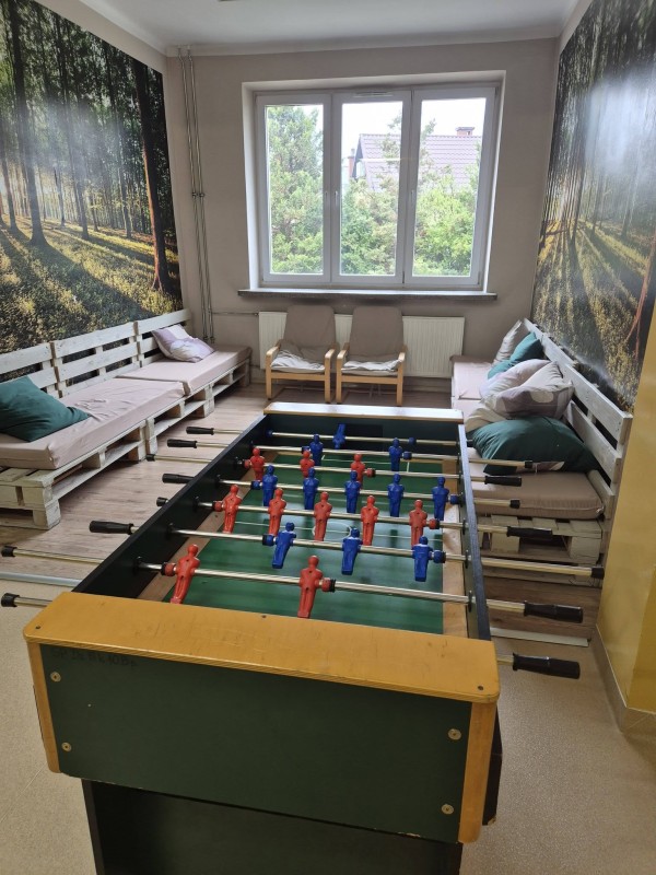 asna strefa relaksu z dużym stołem do piłkarzyków w centrum. Po bokach niskie kanapy z palet i fototapety z motywem lasu. W głębi okno i dwa fotele. Wnętrze przytulne, łączące sportową rozrywkę z naturalnym, nowoczesnym stylem.