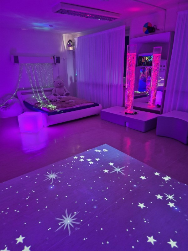 Sali Doświadczania Świata z fioletowym oświetleniem. Na podłodze projekcja gwiazd, obok łóżko z kurtyną światłowodów i dwa podświetlone kolumny wodne przed lustrem. Nowoczesna przestrzeja sensoryczna służąca relaksacji i terapii.