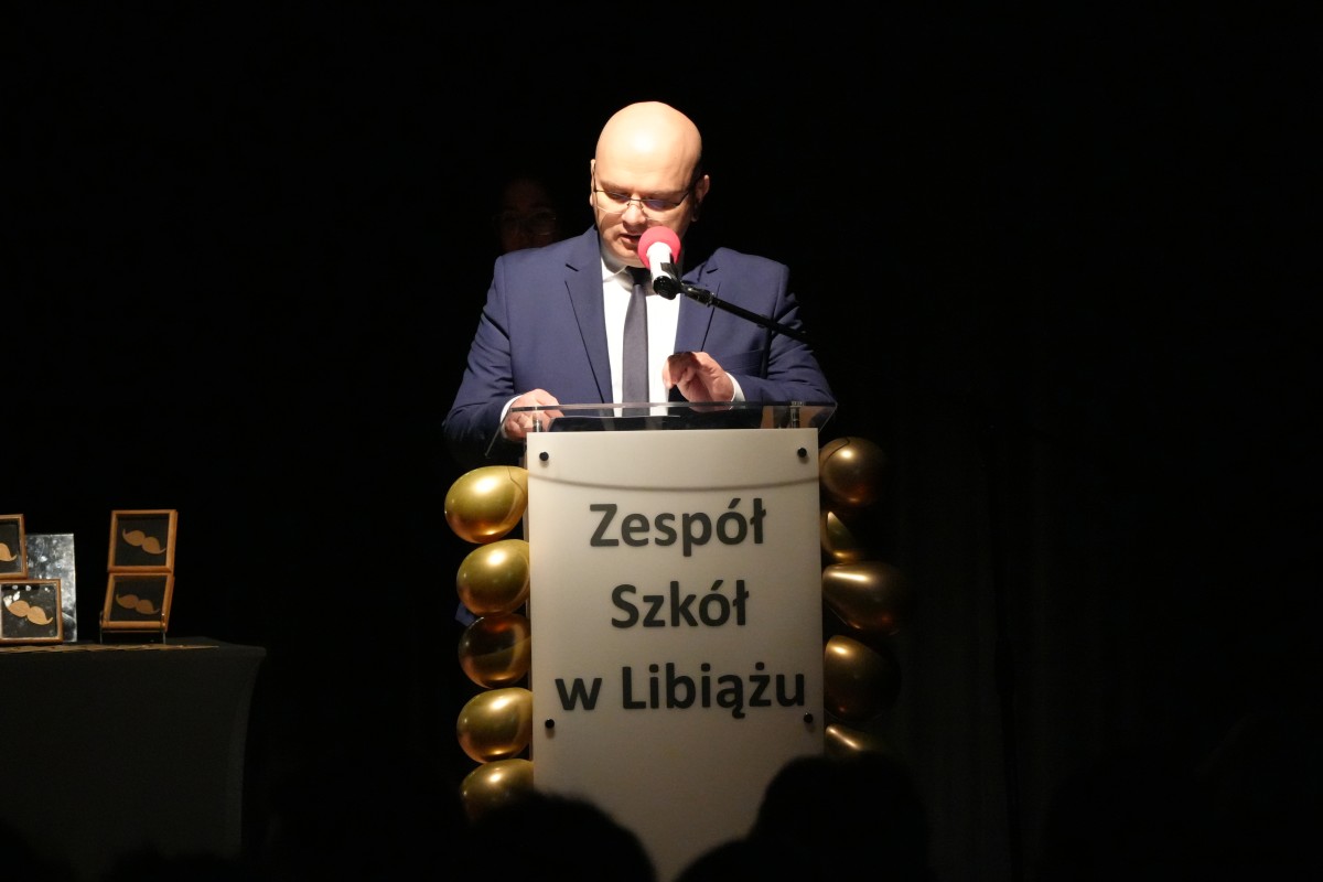 Mężczyzna przemawia zza pulpitu, na którym napisana jest nazwa szkoły.