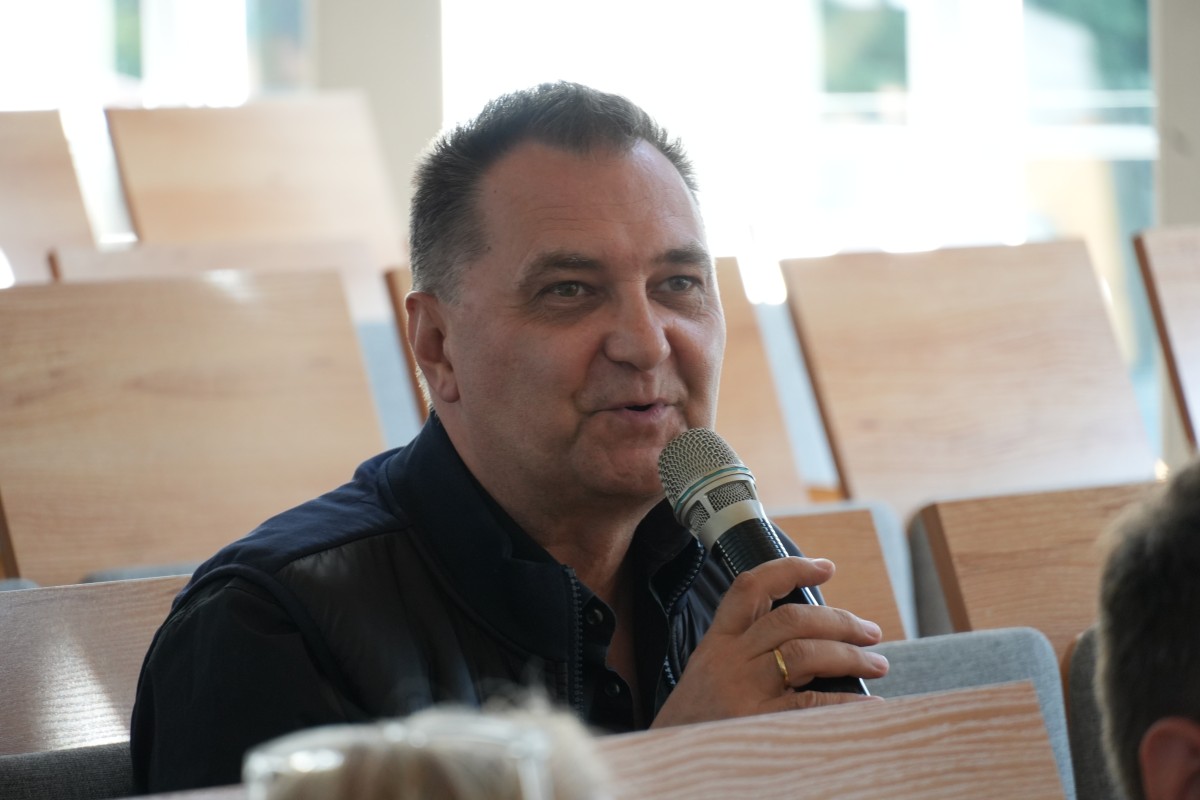 Zdjęcie mężczyzny siedzącego na auli wykładowej. Trzyma w ręce mikrofon i mówi do niego.