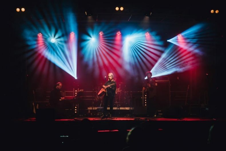 Trzech muzyków na scenie oświetlonej w kolorze czerwono-niebieskim . Dwóch grających na gitarach jeden na klawiszach. 