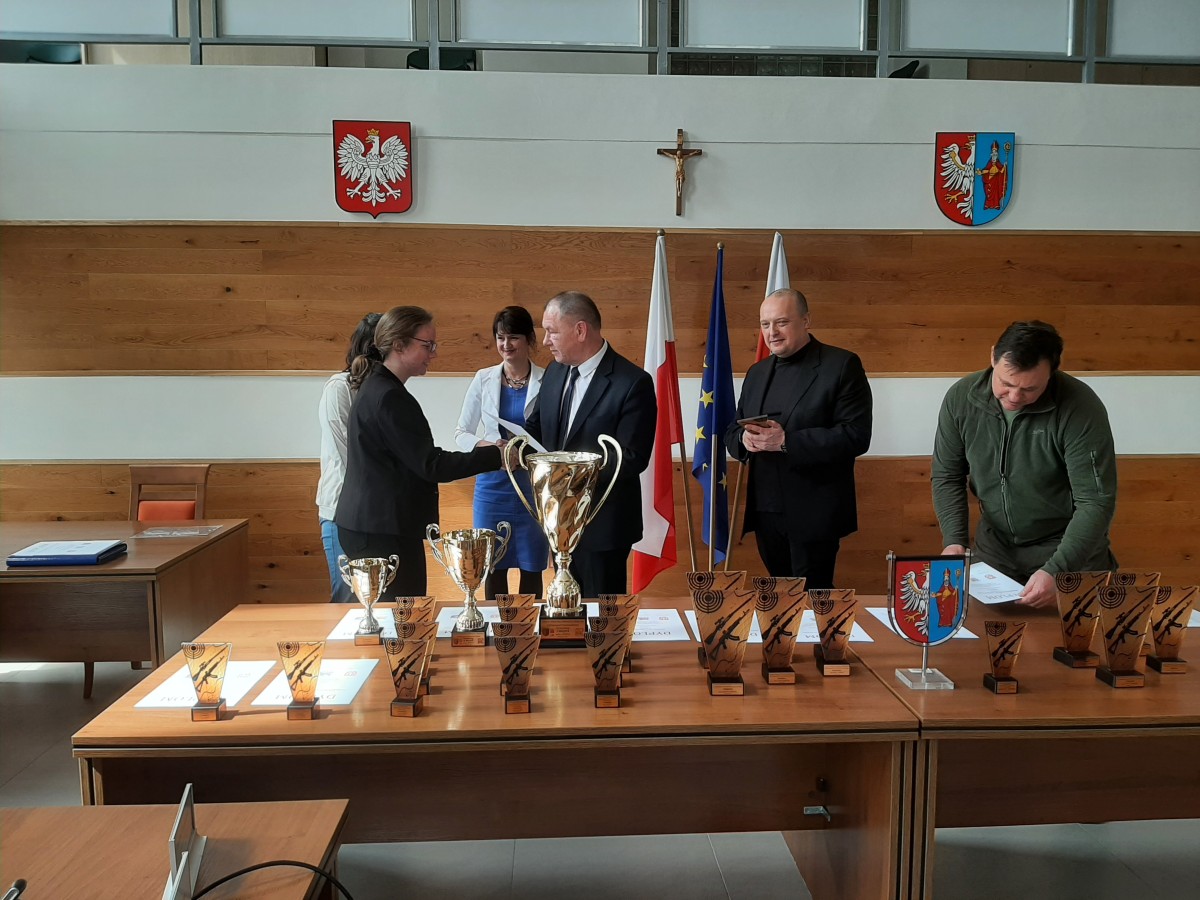 Trzy kobiety i trzecz mężczyzn eleganco ubranych w kolorach zielonym , czarnym i niebieskim. Na tle złotych pucharów i statuetek z czarnym nadrukiem. W tyle flaga Polski i UE.