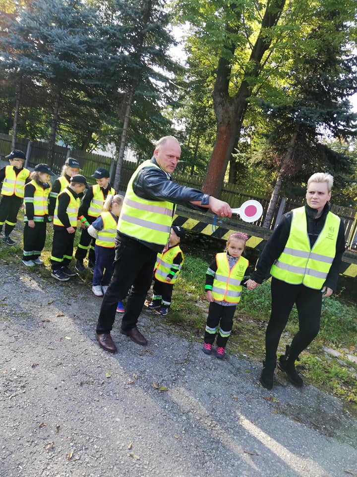 Mężczyzna w odblaskowej kamizelce wskazuje  lizakiem kierunek dzieciom w mundurach strażackich i kamizelkach odblaskowych 