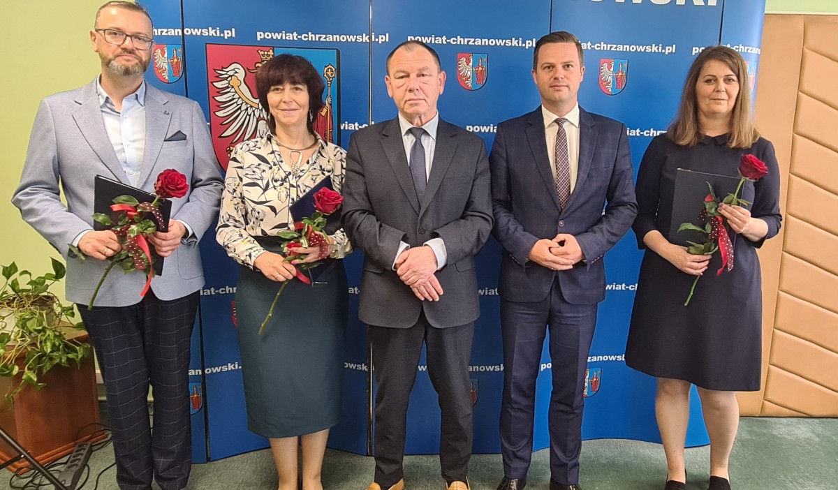 Trzech mężczyzn i dwie kobiety stoją na tle niebieskiej ścianki z białymi napisami i herbem w kolorze czerwono, żółto niebiesko białym w czarnej ramce