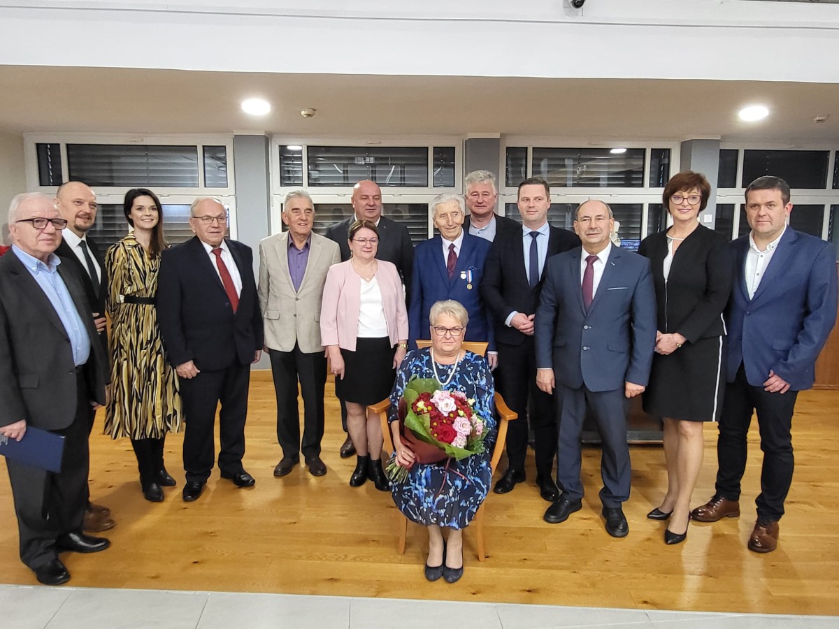 Kobieta siedzi na stołku, za nią rząd osób 
