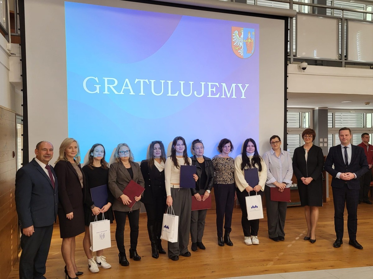 Zdjęcie władz powiatu, uczennic, rodziców, nauczycieli i dyrekcji szkół. Na zdjęciu znajduje się 12 osób. W tyle, na rzutniku wyświetlony jest napis GRATULUJEMY oraz herb powiatu Chrzanowskiego.