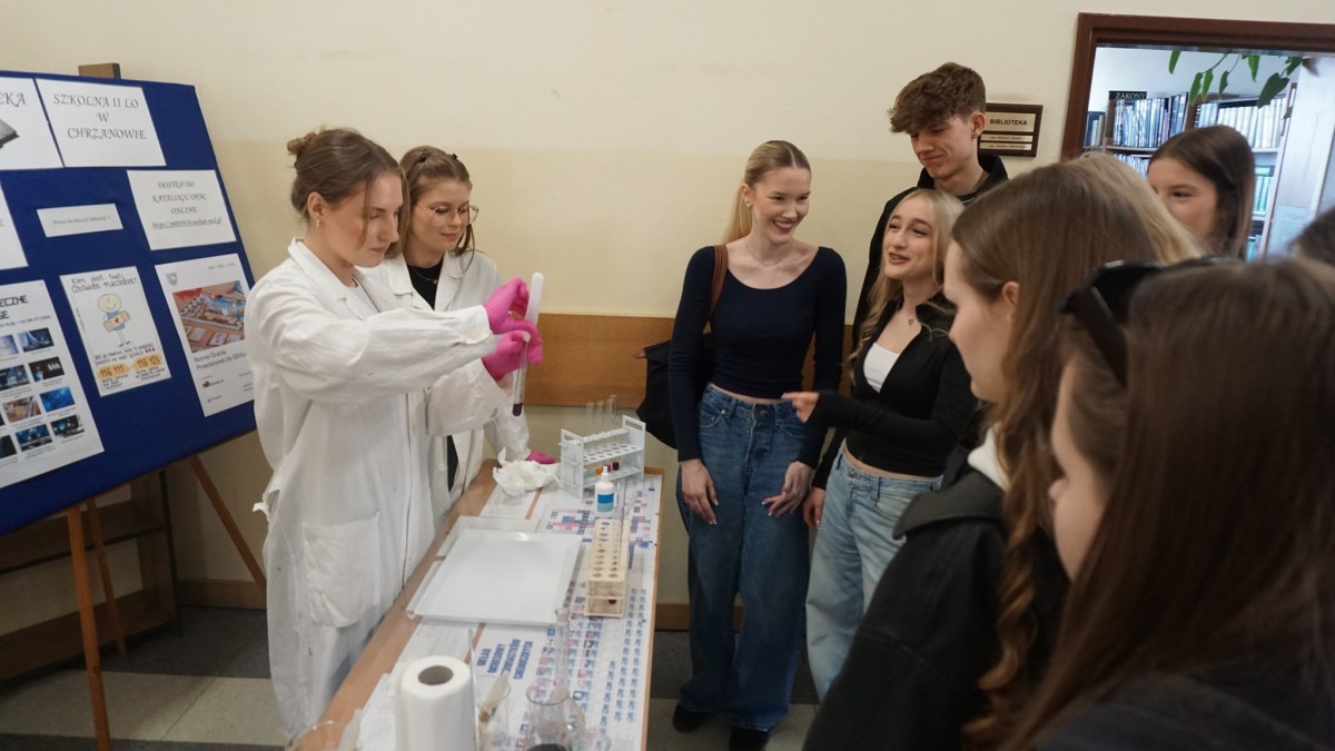 Dwie uczennice w białych fartuchach wykonują eksperymenty chemiczne przed grupą osób 
