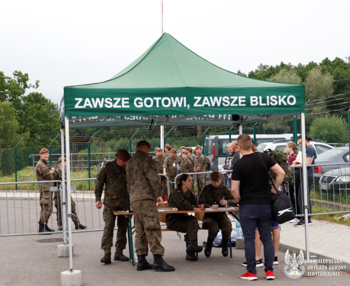 Grupa ludzi pod zadaszeniem, w tle żółnierze