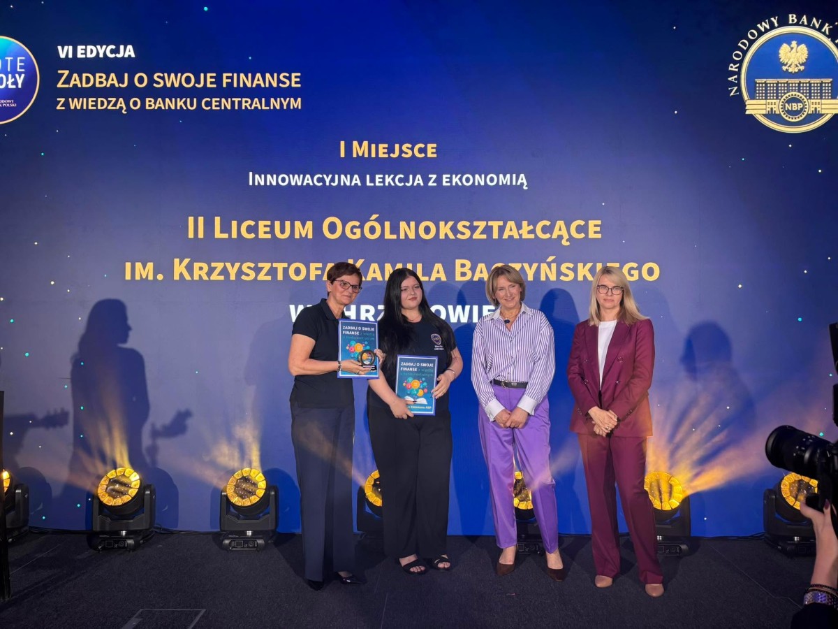 Kobiety stoją na tle tablicy informującej o konkursie