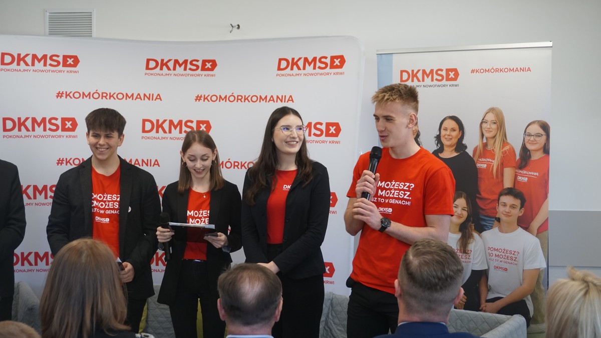 Zdjęcie 4 młodych osób. Za nimi roll'upy i ścianki promocyjne. Jeden z uczniów trzyma mikrofon i przemawa do grupy osób.