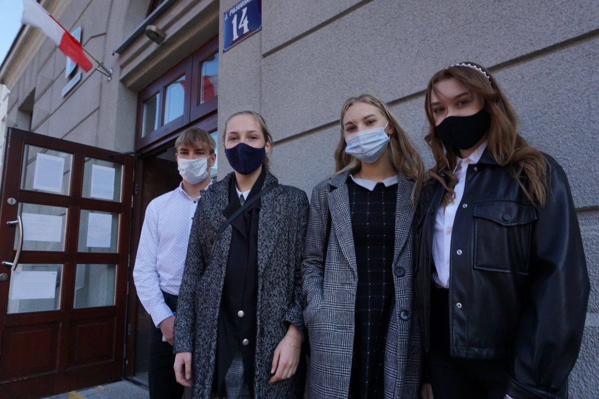 Chłopiec w białej koszuli stoi obok trzech dziewcząt. Wszyscy są w maseczkach. Za nimi budynek szkoły. 