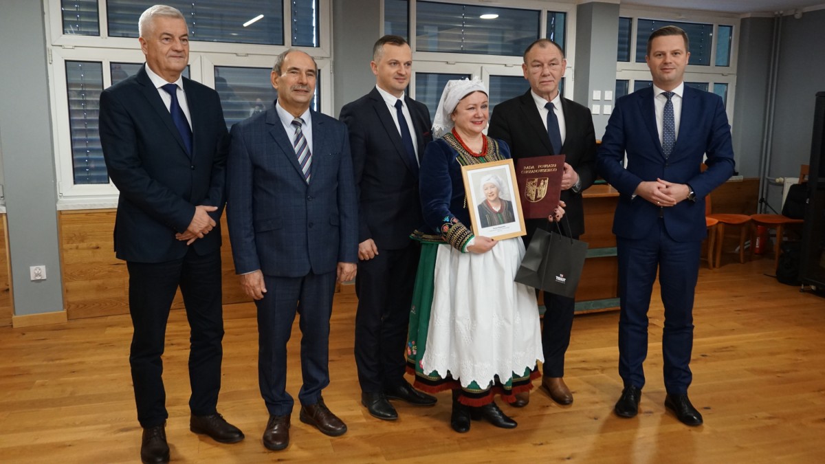 Kobieta w kolorowym stroju regionalnym. Na głowie czepiec . Obok mężczyźni w garniturach 