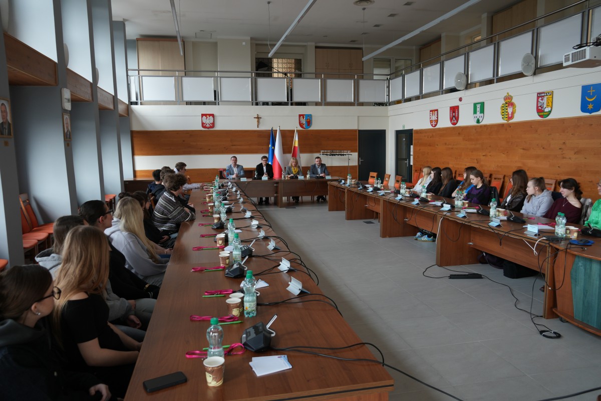 Zdjęcie zebranych uczniów siedzących na sali narad. Przy stole prezydialnym zasiadają przedstawiciele Starostwa Powiatowego.