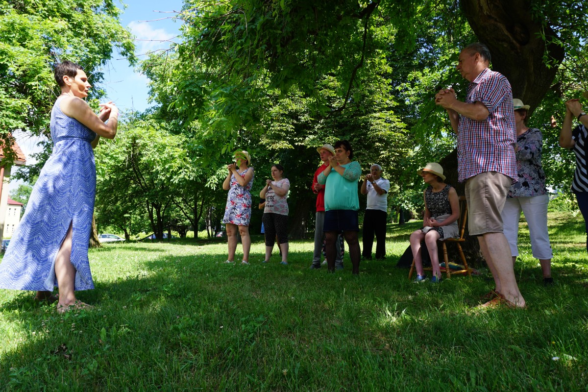 Zajęcia ruchowe w plenerze, prowadzi kobieta w zwiewnej sukni. Przed nią kobiety i mężczyźni 