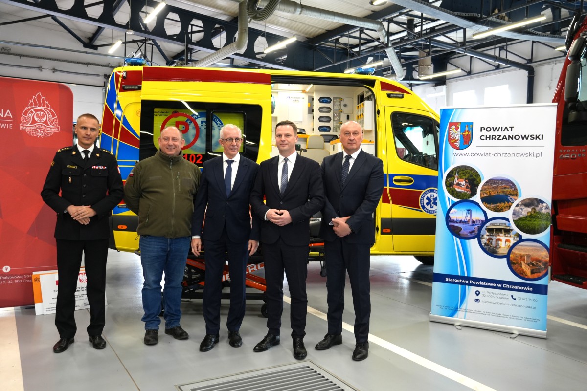 Pięciu mężczyzn pozuje do zdjęcia. W tle karetka, banery promocyjne