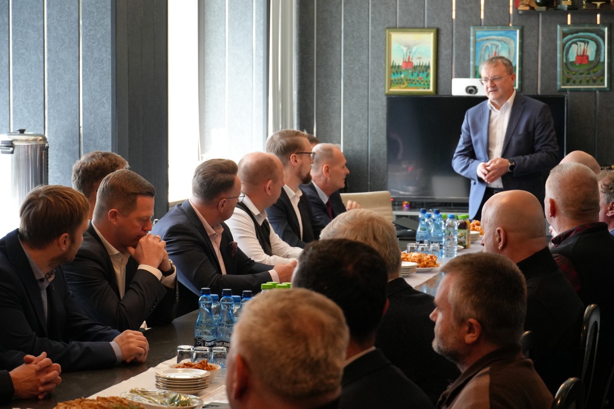 Zdjęcie kilkudziesieciu osób siedzących przy prostokątym stole. Jedna z osób, na szczycie stołu stoi i mówi do zebranych. 