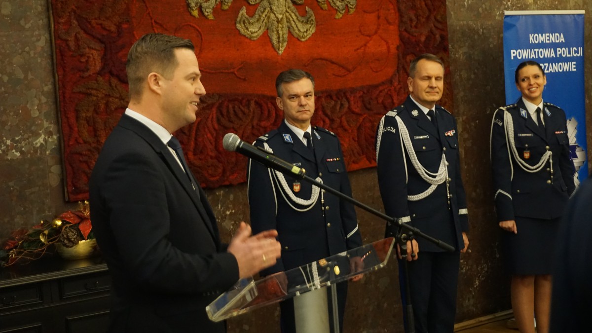 Zdjęcie czterech osób. Trzy osoby stoją w mundurach, jedna w graniturze za mównicą.