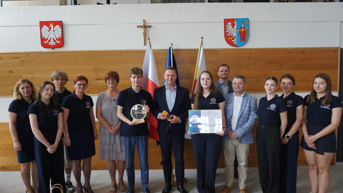 Zdjęcie trzynastu osób w różnym wieku i różnej płci. Kilka osób trzyma statuetki i dyplomy. W tle znajdują się trzy flagi, dwa herby oraz krzyż.
