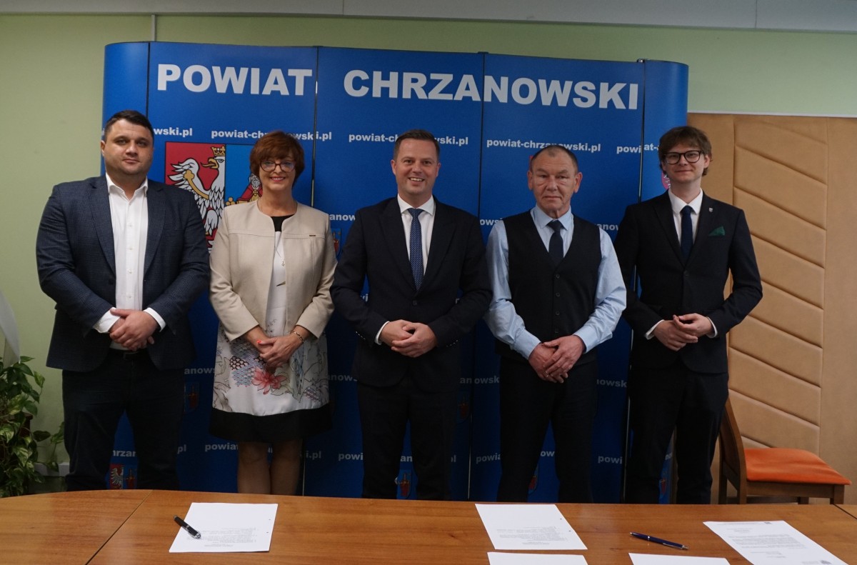 zdjęcie portretowe , członkowie zarządu powiatu chrzanowskiego stojją obok siebie , w tle scianka niebieska z herbami powiatu 