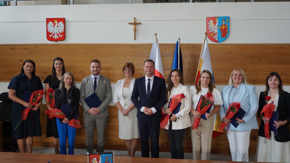 Zdjęcie dziesięciu osób stojących obok siebie, ustawionych do wspólnego zdjęcia. Na zdjęciu osiem kboeit i dwóch mężczyzn. Wszyscy elegancko ubrani. W tyle 2 tarcze w herbami, 3 flagi i krzyż.