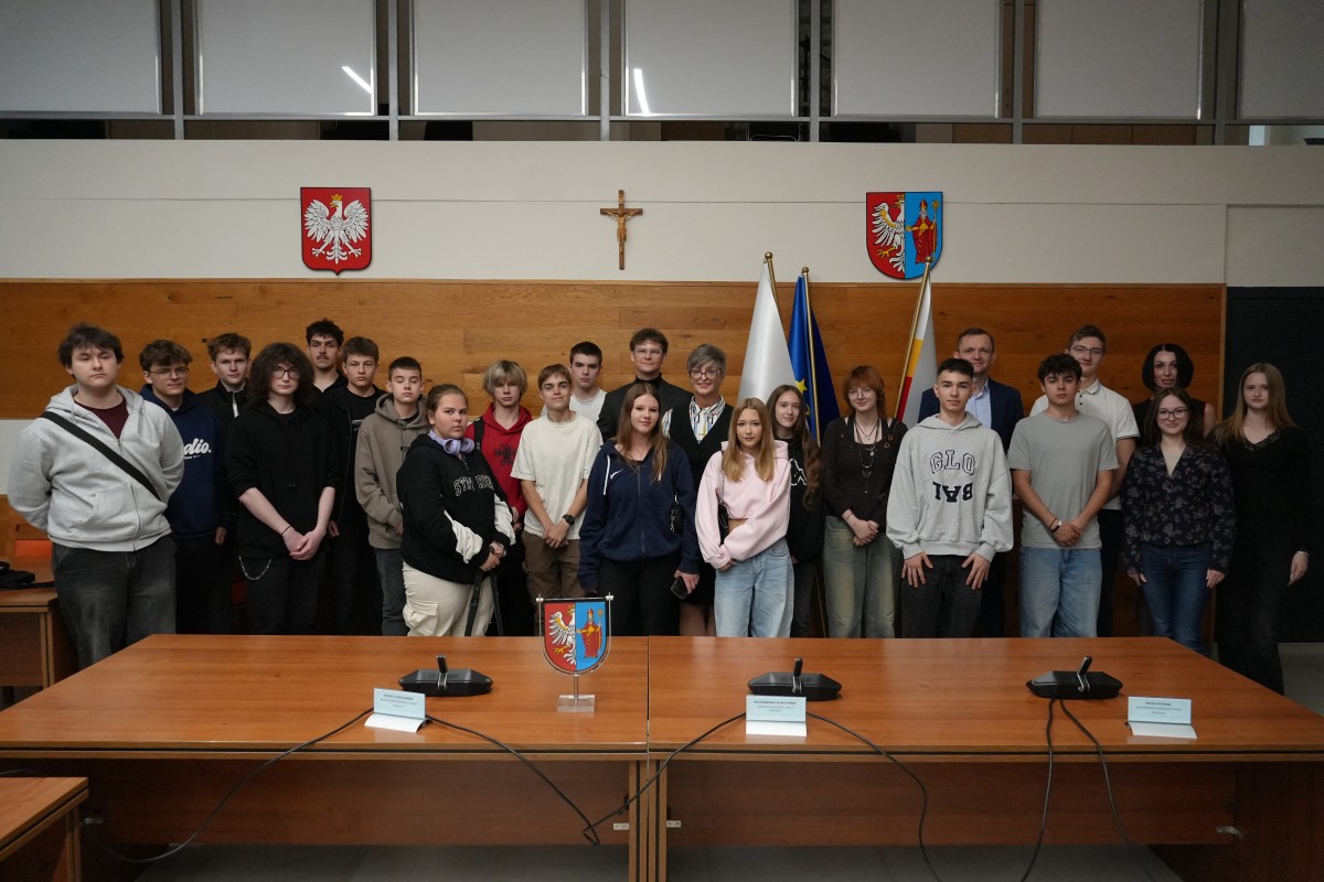 Zdjęcie kilkudziesięciu uczniów wraz z trzema przedstawicielami urzędu. W tle herb powiatu, godło państwa oraz krzyż.