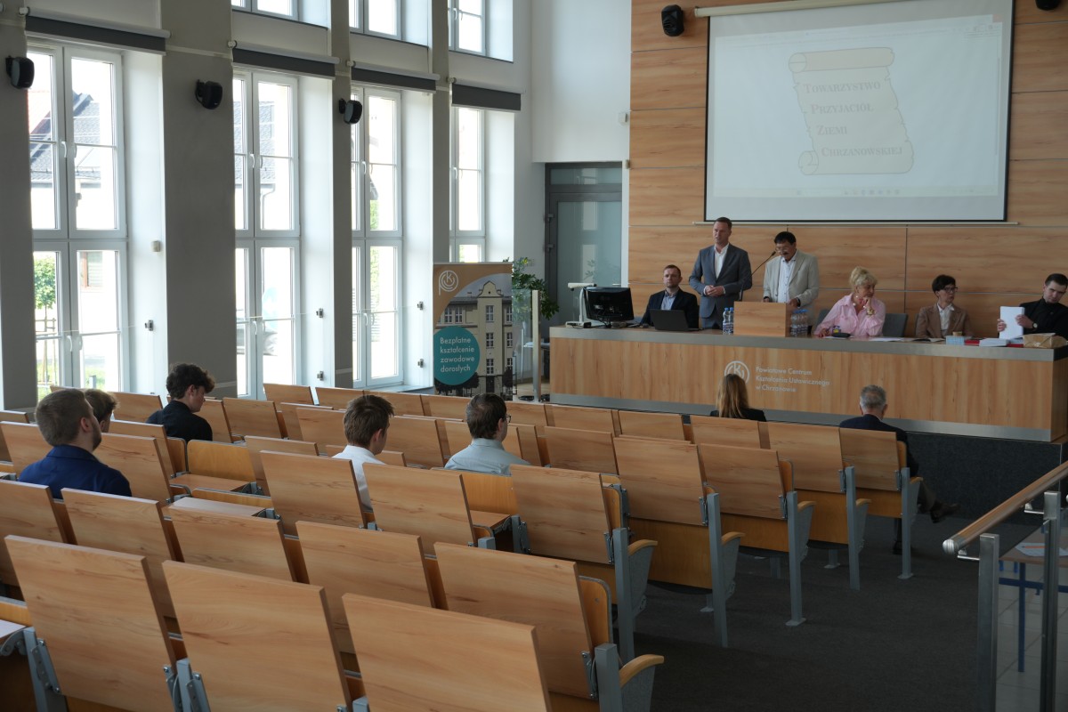 Zdjęcie kilku uczniów siedzących na sali wykładowej i patrzącej w stole podium, na którym siedzi i stoi kilak dorosłych osób. 
