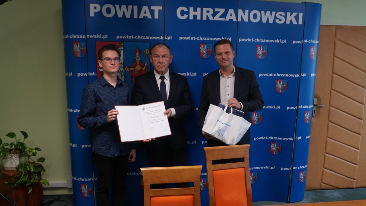 Trzech mężczyzn na tle niebieskiej ścianki z białymi napisami i herbem w kolorze biało, czerwono, żółtym