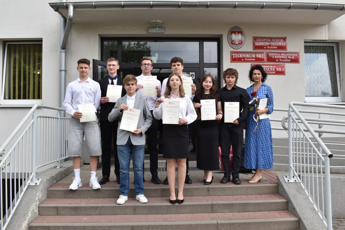 NA ZDJĘCIU 9 OSÓB, UCZNIOWIE ZSTU WRAZ Z OPIEKUNEM W NIEBIESKIEJ SUKIENCE  STOJĄ PRZED SZKOŁĄ  NA SCHODACH, UBRANI W GALOWE STROJE ORAZ PREZENTUJĄ DO ZDJĘCIA DYPLOMY 