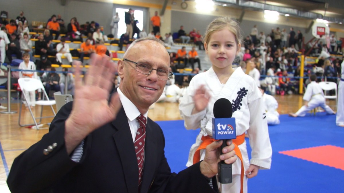 Uśmiechnięty mężczyzna macha ręką do kamery. W drugiej ręce trzyma mikrofon. Obok mała dziewczynka - zawodniczka karate 