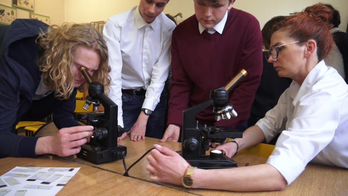 Nastolatek patrzy przez mikroskop. Obok nauczycielka i inni chłopcy. 