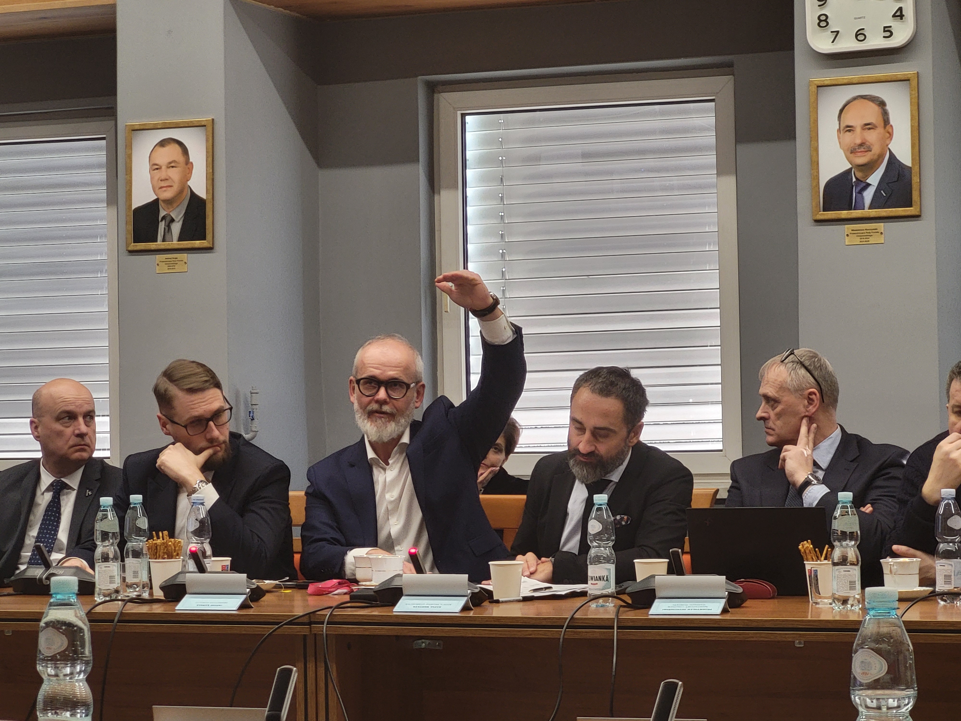 Zdjęcie kilku mężczyzn siedzacych przy stole na sali konferencyjnej. Jeden z nich gestykuluje podczas wypowiedzi. 