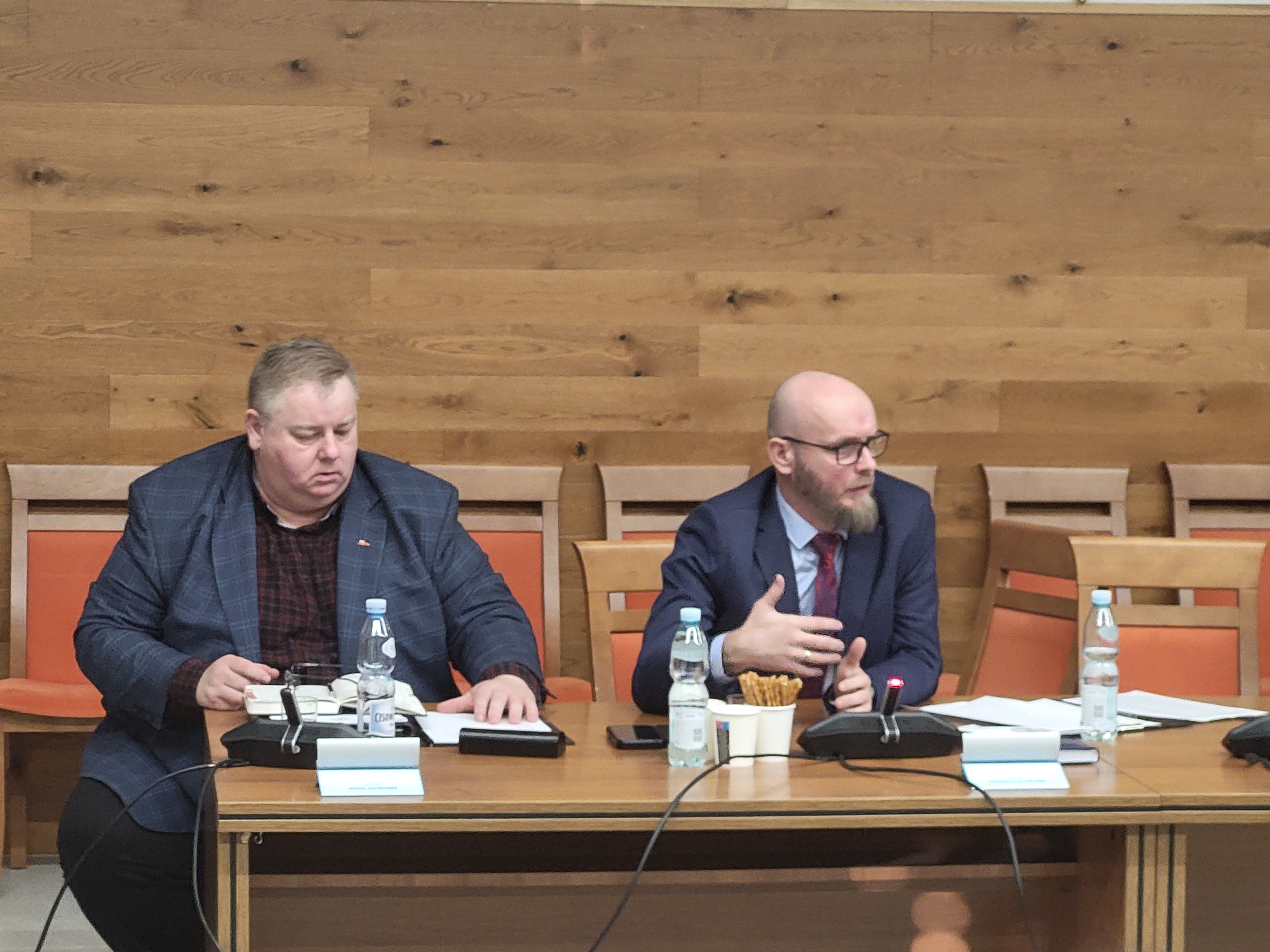 Zdjęcie dwóch mężczyzn siedzacych przy stole na sali konferencyjnej. Jeden z nich gestykuluje podczas wypowiedzi. 