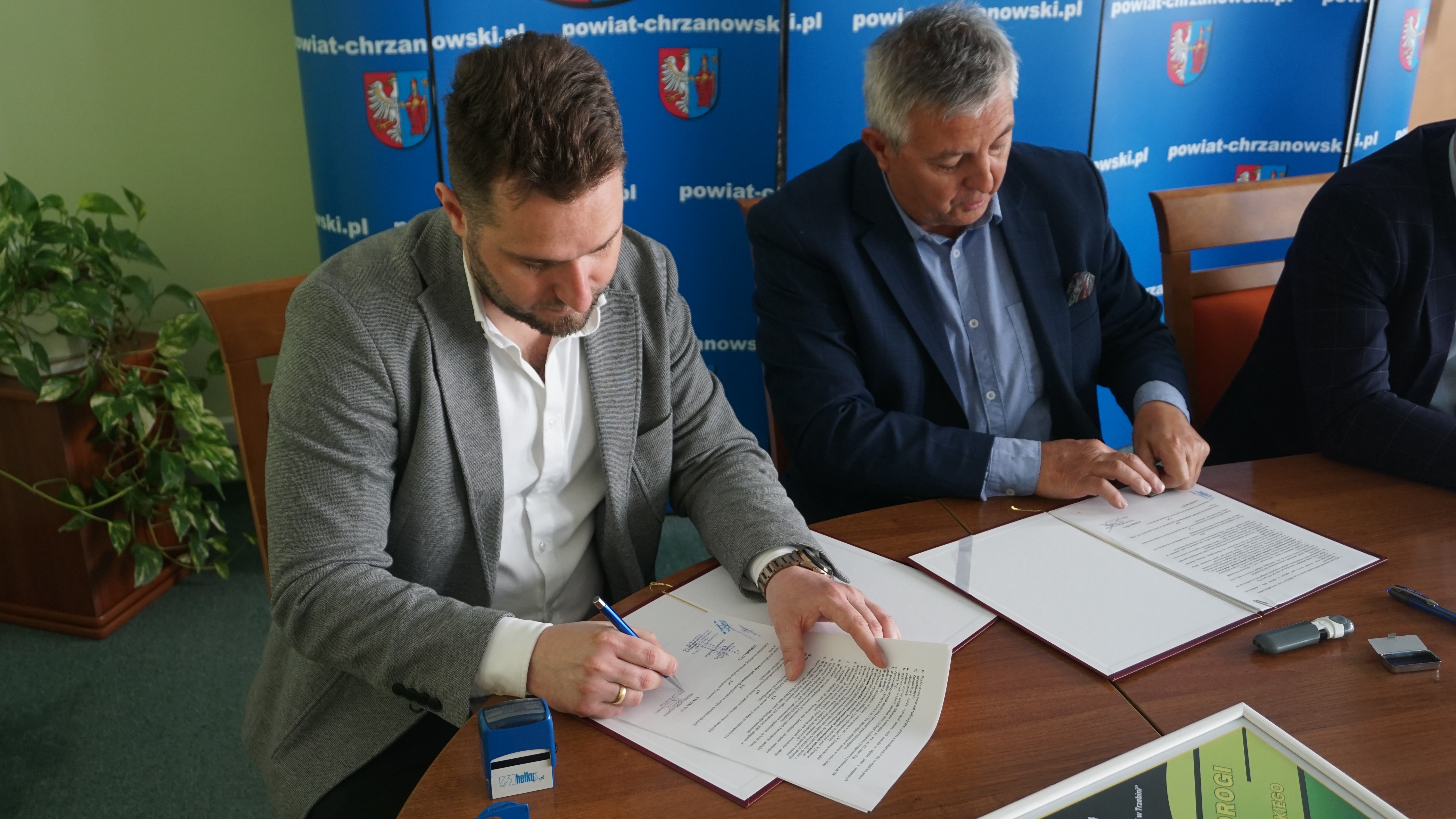 Zdjęcie dwóch mężczyzn siedzących przy stole. Oboje podpisują dokumenty. 