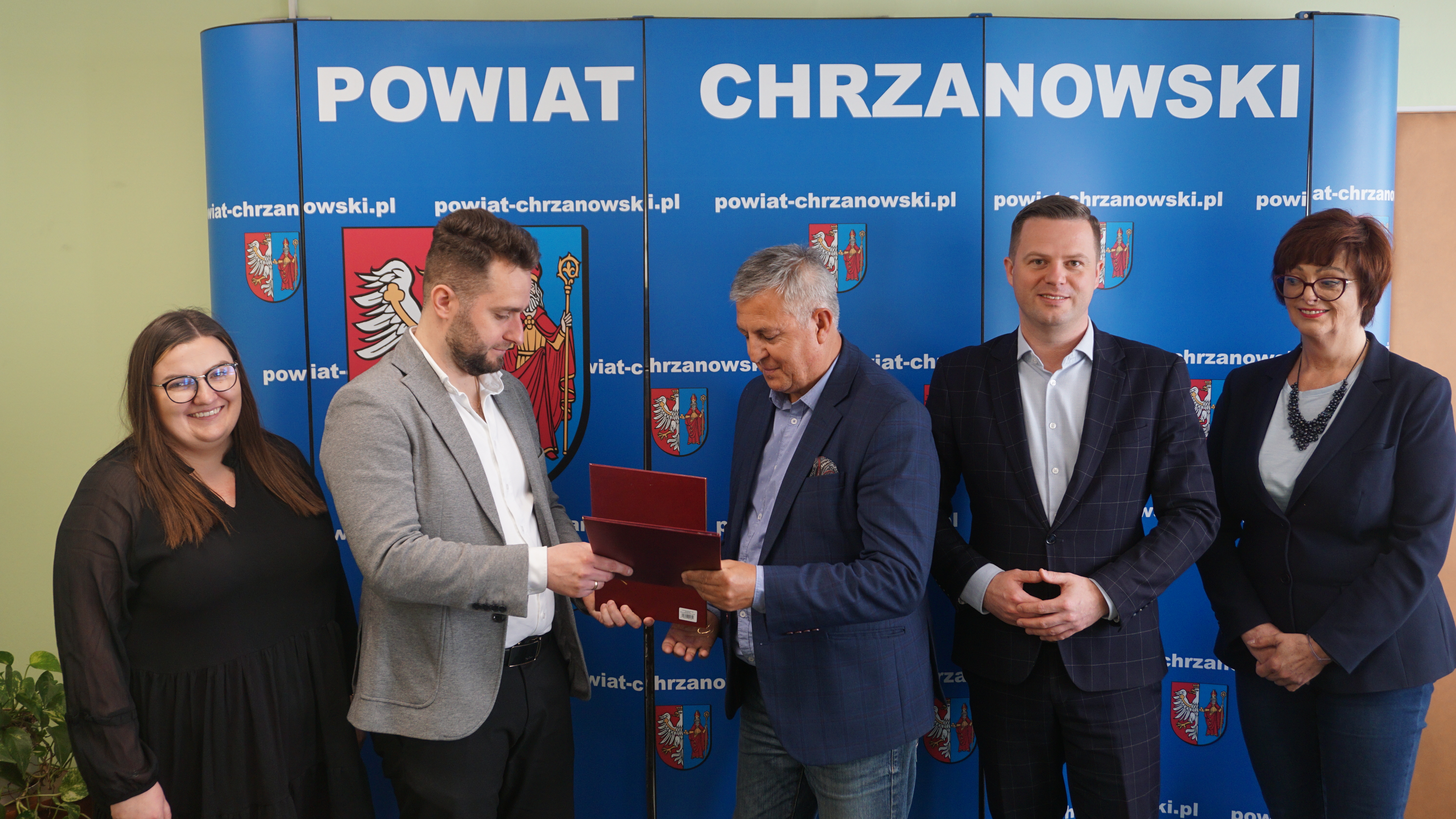 Zdjęcie 5 osób stojących przy ściance promocyjne Powiatu Chrzanowskiego. Dwójka z nich wymienia się teczkami. Od lewej: dwójka przedstawicieli firmy WIKOS FRESH, dyrektor PZD, starosta oraz wicestarosta chrzanowski.