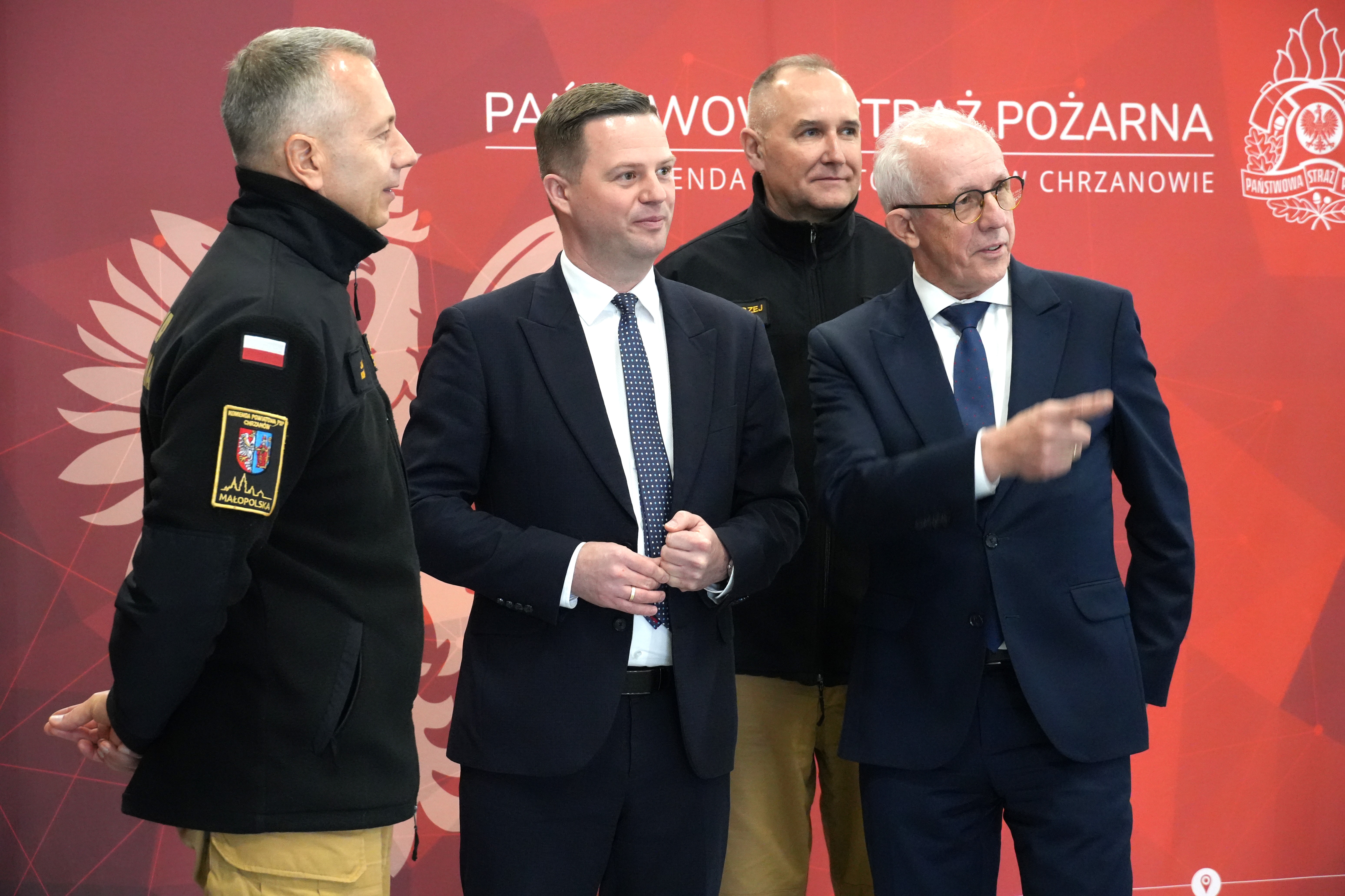 Kilku mężczyzn, jeden  wskazuje ręką na lewo. W tle baner promocyjny. 