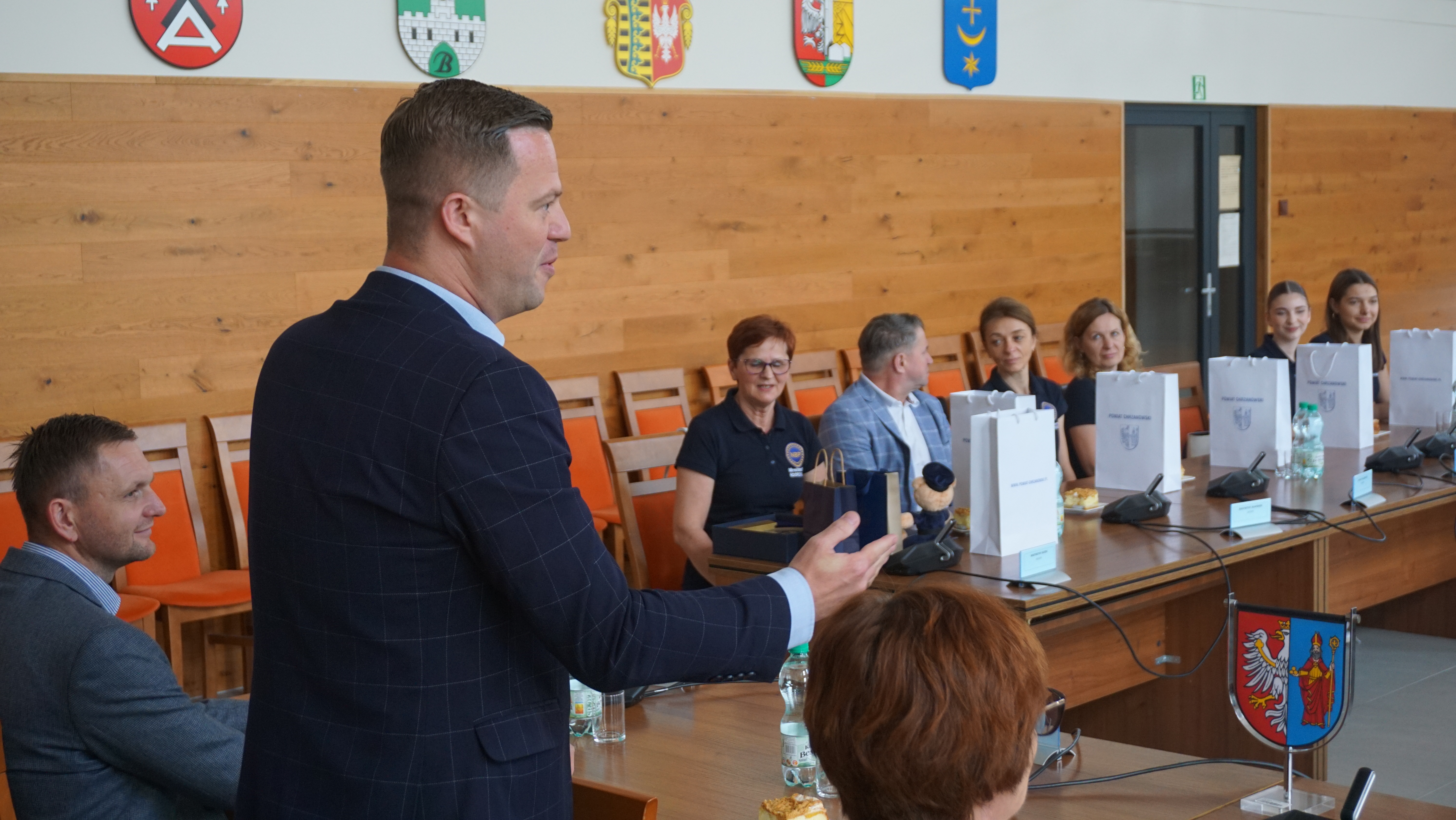 Zdjęcie kilku osób siedzących przy stółach. Starosta stoi mówi do zebranych i gestykuluje. 