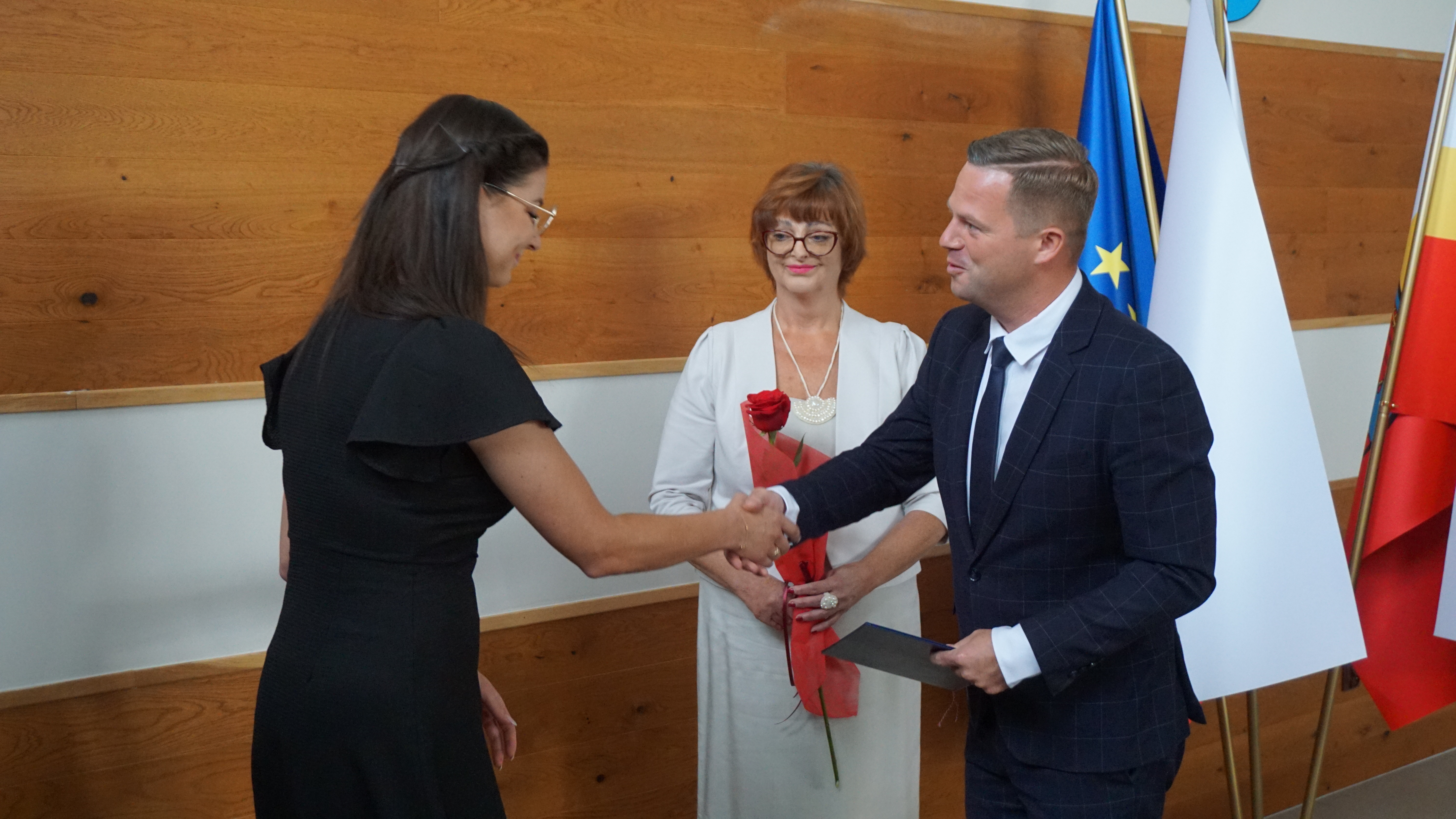 Zdjęcie trzech osób. Mężczyzna ściska dłoń kobiecie. Druga kobieta w tle się uśmiecha trzymając róże. 