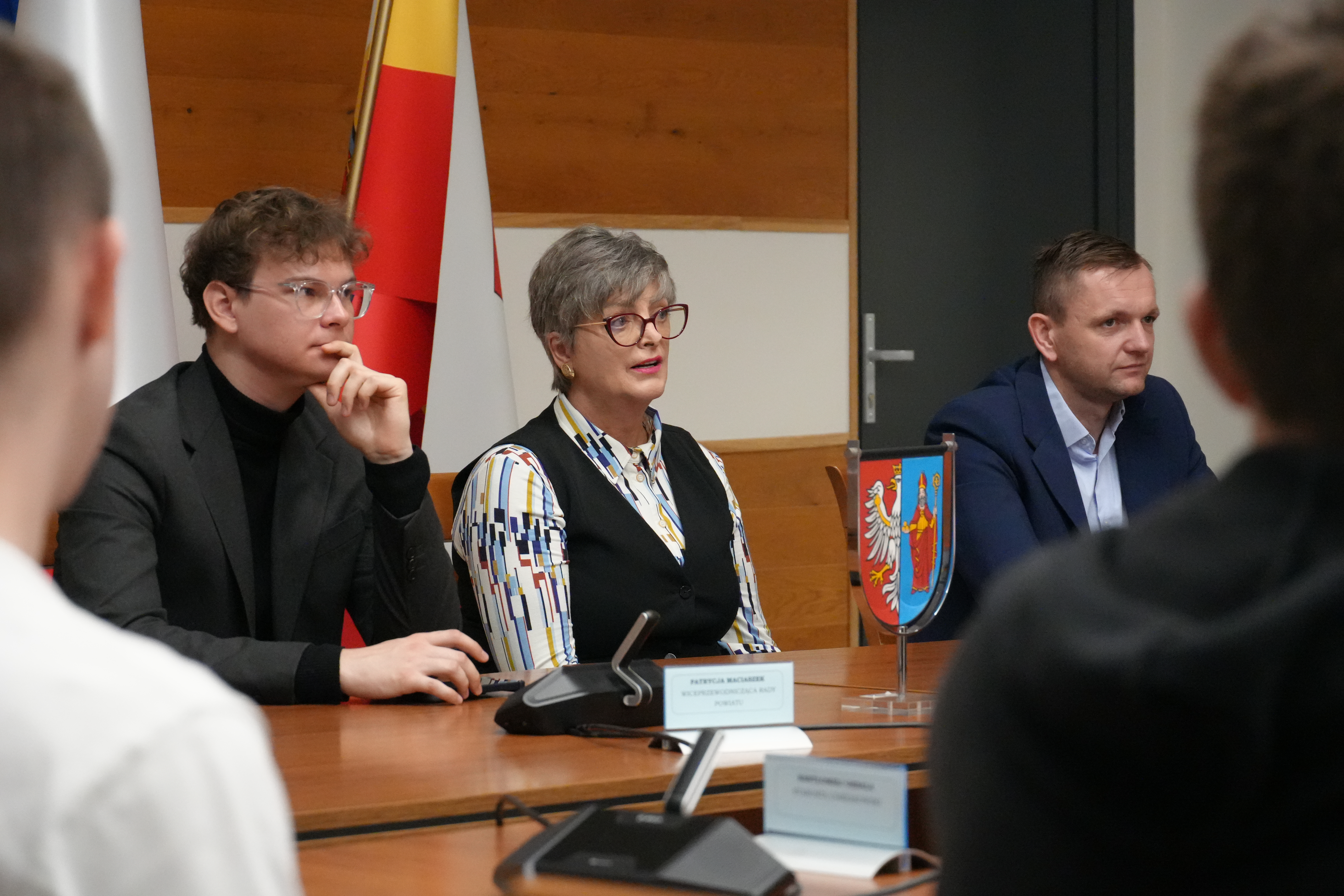 Zdjęcie trzech osób siedzących przy stole. Kobieta w środku mówi do zebranych.