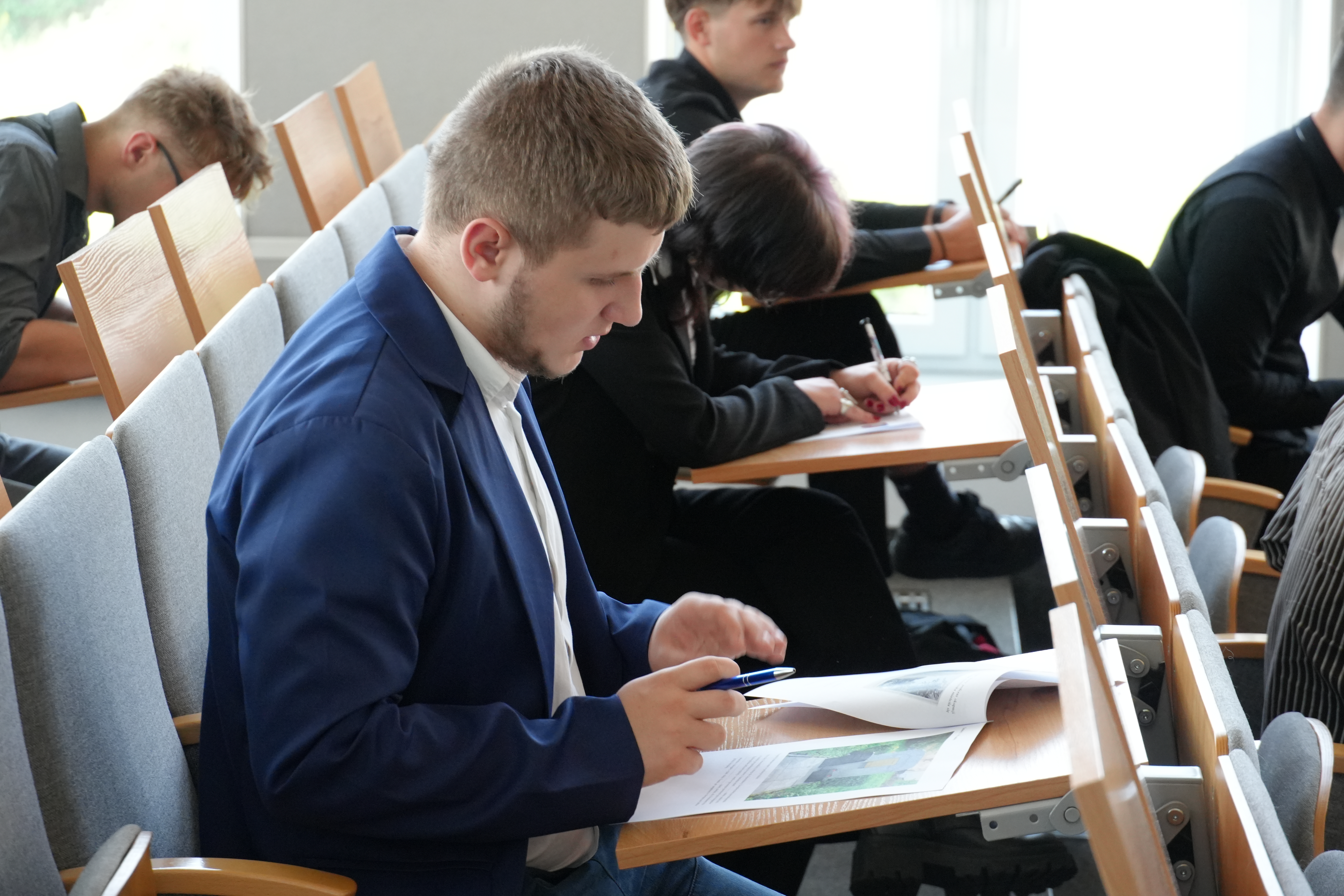 Zdjęcie kilku uczniów siedzących w jednym rzędzie, w oddaleniu od siebie i piszących po kartkach papieru leżących przed nimi
