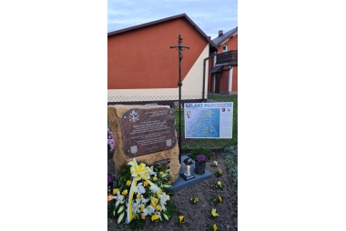 Kamień z tablicą, obok wiązanka kwiatów i znicz 