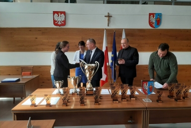 Trzy kobiety i trzecz mężczyzn eleganco ubranych w kolorach zielonym , czarnym i niebieskim. Na tle złotych pucharów i statuetek z czarnym nadrukiem. W tyle flaga Polski i UE.