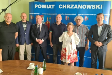 7 osób (1 kobieta,6 mężczyzn) stojących na tle niebieskiego baneru z białym napisemi herbem biało żółto niebieski w czarnej ramce