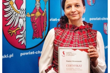 Kobieta trzyma w rękach dyplom. W tle ścianka reklamowa powiatu 