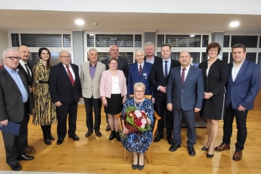 Kobieta siedzi na stołku, za nią rząd osób 