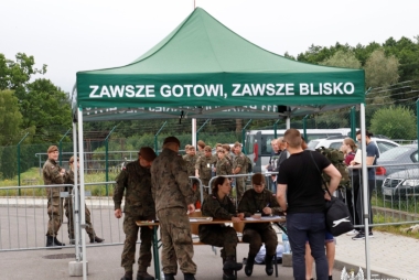 Grupa ludzi pod zadaszeniem, w tle żółnierze