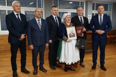 Kobieta w kolorowym stroju regionalnym. Na głowie czepiec . Obok mężczyźni w garniturach 